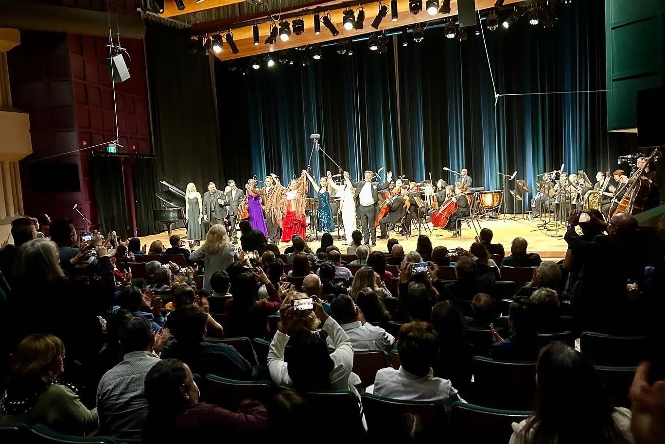 Performers and orchestra receiving applause from an audience in a theatre.